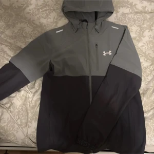 Grå och svart jacka från Under Armour - Säljer en snygg grå och svart jacka från Under Armour. Jackan har en dragkedja framtill och en huva för extra skydd. Perfekt för kyligare dagar med sin stilrena design och funktionella detaljer.
