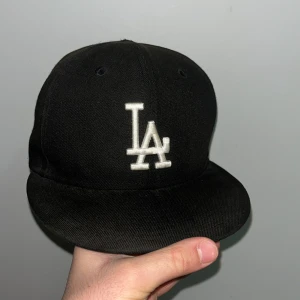 Svart keps med LA-logga - Snygg svart keps från New Era med vit LA-logga framtill. Kepsen har en rak skärm och är perfekt för en casual look. Den har även en liten MLB-logga baktill. Kepsen är använd en del och därför det kommer priset att sänkas den köptes för 499 men kommer säljas för 250 för att den är använd och såklart kan priset diskuteras.