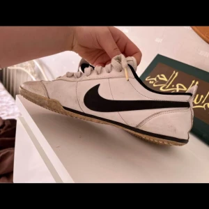 Vita Nike sneakers med svart swoosh - Säljer ett par vita Nike sneakers med den klassiska svarta swoosh-loggan på sidan. Skorna har snörning och en bekväm passform. De är lite smutsiga på sulan men annars i bra skick. Perfekta för vardagsbruk eller träning!