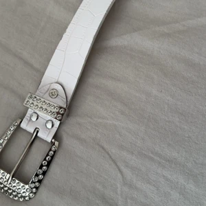 Vitt glittrigt bälte - Säljer ett supercoolt vitt bälte med glittriga detaljer. Bältet har en krokodilpräglad yta och är dekorerat med massor av bling i form av strass. Spännet är också täckt med strass, vilket ger en extra touch av glamour. Perfekt för att liva upp vilken outfit som helst!