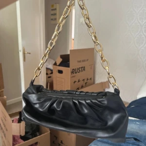 Svart axelväska med guldkedja - Säljer en snygg svart axelväska i skinn med en cool guldkedja som axelrem. Väskan har en rynkad design och är perfekt för både vardag och fest. Den är i nyskick och redo att användas direkt!