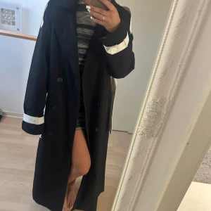 Svart kappa med vita detaljer - Säljer en snygg svart kappa med vita detaljer på ärmarna. Den har en klassisk design med knappar framtill och en elegant krage. Perfekt för höst och vår!