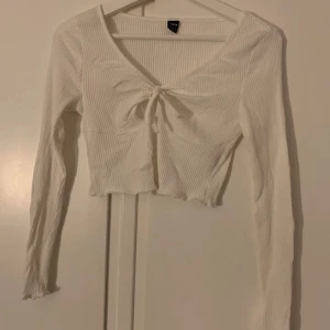 Vit ribbad topp från Shein - Säljer en vit ribbad topp från Shein med lång ärm och knytning framtill. Toppen är croppad och har en v-ringad design som ger en stilren look. Perfekt för att matcha med höga jeans eller kjol.
