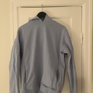 Ljusblå hoodie från Sandro - Säljer en stilren ljusblå hoodie från Sandro. Tröjan har en klassisk design med långa ärmar och en bekväm passform. Perfekt för en avslappnad look.