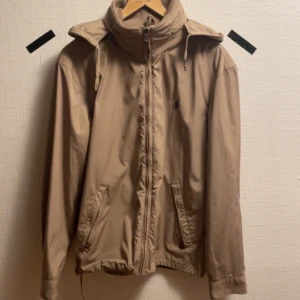 Beige jacka från Polo Ralph Lauren - Säljer en stilren beige jacka från Polo Ralph Lauren. Jackan har dragkedja framtill och en hög krage med justerbar huva. Perfekt för svala dagar. Storlek S-M. Inga defekter. Pris kan diskuteras 