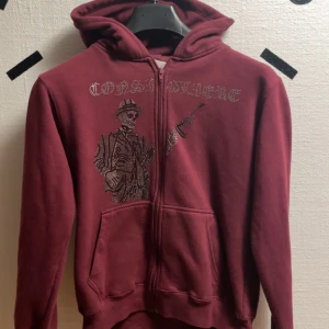 Vinröd hoodie från Costino Stockholm - Snygg vinröd hoodie från Costino Stockholm med dragkedja och huva. Framsidan pryds av ett coolt tryck med texten 'Consigliere' och en bild av en man med gevär. Riktigt fet om du frågar mig. Pris kan diskuteras 