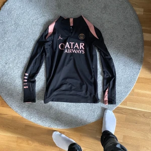 Svart långärmad träningströja från Jordan - Säljer en svart långärmad träningströja från Jordan med rosa detaljer och dragkedja vid halsen. Tröjan har tryck med 'Qatar Airways' och PSG-logga på bröstet. Perfekt för fotbollsträning eller annan sportaktivitet. strlk M