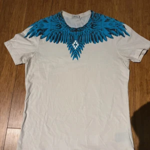 Vit t-shirt med blått mönster från Marcelo Burlon - Säljer en vit t-shirt från Marcelo Burlon med ett coolt blått mönster runt halsen. T-shirten har korta ärmar och är perfekt för en avslappnad stil. Märket är känt för sina unika och trendiga designer.