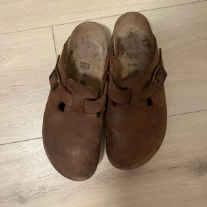 Bruna birkenstocks med spänne - Säljer ett par bruna birkenstocks med spänne. De har en enkel och stilren design med en öppen häl och rundad tå. Perfekta för en avslappnad stil.