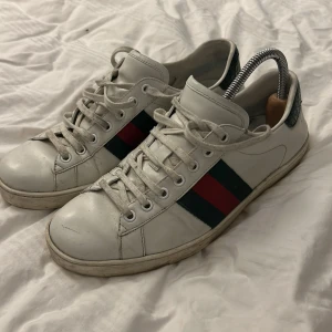 Vita Gucci sneakers med ränder - Snygga Gucci sneakers med klassiska gröna och röda ränder på sidan. Skorna har snörning och en låg profil, perfekt för en stilren look. De har en vit sula och detaljer i svart på hälen.