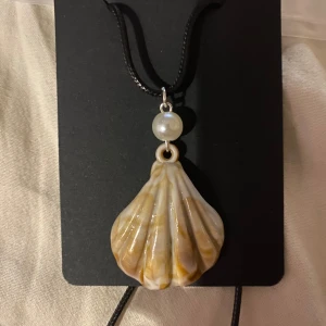 Halsband med snäckformad berlock - Egengjort halsband med en snäckformad berlock i naturfärger och en vit pärla ovanför. Hängs på en svart rem. Perfekt för att ge en strandig känsla till din outfit.