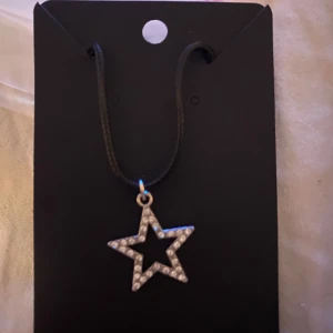Halsband med stjärnhänge - Egengjort halsband med ett stjärnformat hänge dekorerat med glittrande stenar. Hänget hänger på en svart rem och ger en stilren look. Perfekt för att lägga till lite bling till din outfit.