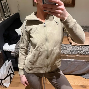 Grön hoodie från Peak Performance - Säljer en grön hoodie från Peak Performance med dragkedja och justerbar huva. Tröjan har långa ärmar och en broderad logga på bröstet samt en ficka på ärmen. Perfekt för en avslappnad stil.