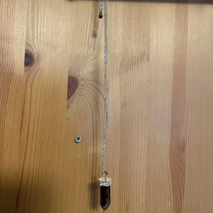 Halsband med lila kristallhänge - Elegant halsband med en lila kristallformad hänge. Kedjan är i silverfärg och hänget har en dekorativ kant med små stenar. Perfekt för att ge en bohemisk touch till din stil.