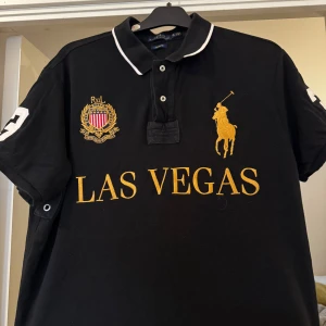 Svart pikétröja från Ralph Lauren - Snygg svart pikétröja från Ralph Lauren med 'Las Vegas' tryck i guld på framsidan.  Perfekt för en stilren look. Sitter som en L Pris kan diskuteras.