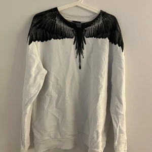 Vit tröja med vingmönster från Marcelo Burlon - Säljer en vit tröja från Marcelo Burlon med ett coolt svart vingmönster som sträcker sig över axlarna och ner på bröstet. Tröjan har långa ärmar och en rund halsringning. Perfekt för dig som vill ha en unik stil!