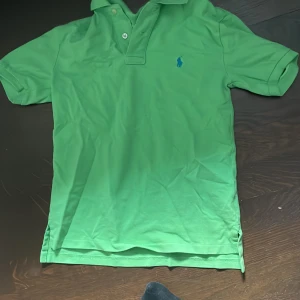 Grön pikétröja från Ralph Lauren - Säljer en snygg grön pikétröja från Ralph Lauren med klassisk krage och knappar. Tröjan har en broderad logotyp i blått på bröstet. Perfekt för en stilren och avslappnad look.