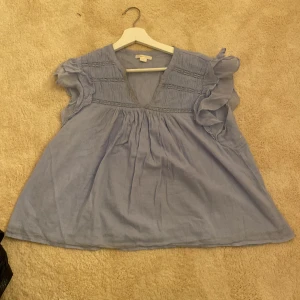 Blå blus från Gina Tricot - Säljer en söt blå blus från Gina Tricot med volangdetaljer på ärmarna och v-ringning. Tröjan har dock sminkfläckar i v ringningen som jag inte testat att ta bort men det går säkert men inte säker, därav lite billigare pris!.