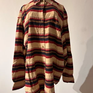 Randig oversized flanellskjorta från H&M - Säljer en cool, randig flanellskjorta från H&M i storlek 40. Skjortan har ett mönster i beige, vinröd och svart -jordnära färger och en ficka på bröstet. Perfekt för en avslappnad stil när det är kyligt.