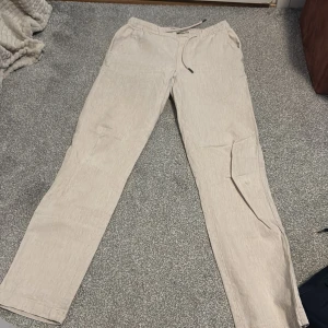 Beige linnebyxor från Dressmann - Snygga beige linnebyxor från Dressmann i storlek XS. Byxorna har en avslappnad passform med snörning i midjan för extra komfort. Perfekta för en stilren och bekväm look.