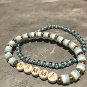 Armband med texten 'MAMMA' - Två armband med pärlor i blått, vitt och guld. Ett av armbanden har bokstavspärlor som bildar ordet 'MAMMA'. Perfekt som en personlig present eller för att bära själv. 100 för båda 50 för ett