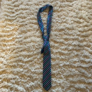 Randig slips från Oscar Jacobson - Snygg randig slips från Oscar Jacobson i blått, grönt och lila. Perfekt för att ge en färgklick till din outfit. Slipsen har en klassisk design med diagonala ränder och är tillverkad i ett elegant material.