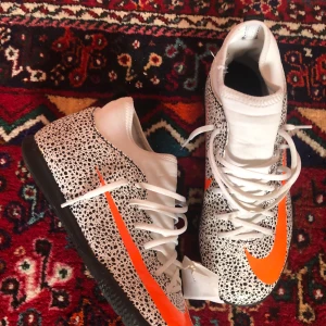 Vita och orangea fotbollsskor från Nike - Snygga fotbollsskor från Nike med ett unikt prickigt mönster i svart och vitt. Skorna har en iögonfallande orange swoosh och snörning för bra passform. Perfekta för inomhusfotboll eller konstgräs.