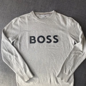 Grå stickad tröja från BOSS - Säljer en stilren grå tröja från BOSS med svart logotyp på bröstet. Tröjan har långa ärmar och ribbade muddar vid ärmslut och nederkant. Perfekt för en avslappnad look.