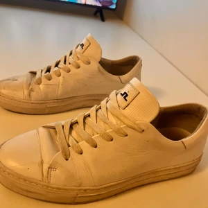 Vita sneakers från J.Lindeberg - Snygga vita sneakers från J.Lindeberg med klassisk design och snörning. Skorna har en stilren look med märkets logotyp på plösen och hälen. Perfekta för en avslappnad stil.