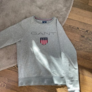 Grå sweatshirt från GANT - Snygg grå sweatshirt från GANT med ett broderat emblem på bröstet. Tröjan har rund hals och långa ärmar, perfekt för en avslappnad stil. Klassisk design med ribbade muddar vid ärmslut och nederkant.