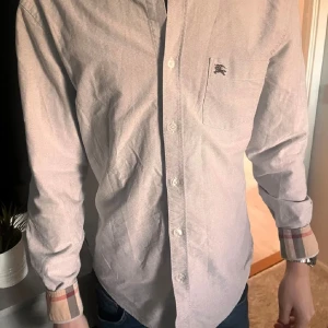 Ljusblå skjorta från Burberry - Snygg ljusblå skjorta från Burberry med klassiska rutiga detaljer på ärmsluten. Skjortan har en broderad logga på bröstfickan och knappar framtill. Perfekt för en stilren look.