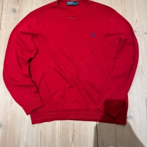 Röd tröja från Polo Ralph Lauren - Säljer en snygg röd tröja från Polo Ralph Lauren med det klassiska blå logotypbroderiet på bröstet. Tröjan har en rund halsringning och långa ärmar, perfekt för en stilren look. Passar utmärkt till både jeans och chinos.