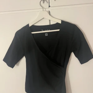 Svart omlottopp från bondelid  - Snygg svart omlottopp från Bondelid i storlek XS. Toppen har korta ärmar och en elegant omlottdesign som ger en stilren look. Perfekt för att klä upp eller ner beroende på tillfälle.