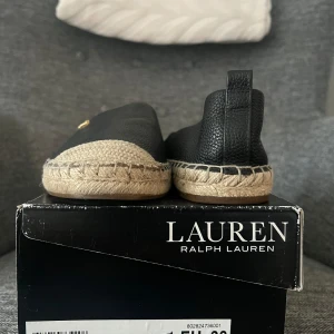 Svarta espadrillos från Ralph Lauren - Snygga svarta espadrillos från Ralph Lauren med flätad sula och diskret logga på ovansidan. Skorna har en klassisk design med en bekväm passform, perfekt för sommarens äventyr.