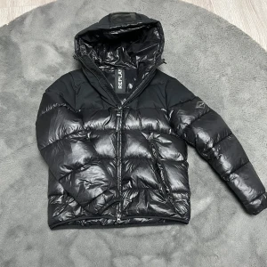 Svart pufferjacka från Replay - Snygg svart pufferjacka från Replay med glansig finish. Jackan har en dragkedja framtill och en huva för extra skydd mot väder. Perfekt för kyligare dagar.