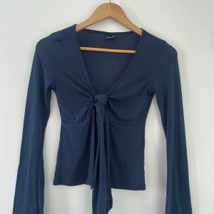 Mörkblå knyttröja från Gina Tricot - Snygg mörkblå tröja från Gina Tricot med knytning framtill. Tröjan har en v-ringad design och långa ärmar, perfekt för en stilren look. Passar bra till både jeans och kjol. Perfekt för höst och vår!