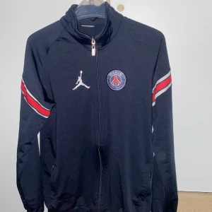 Svart träningsjacka från Jordan x PSG - Snygg svart träningsjacka från Jordan i samarbete med PSG. Jackan har en dragkedja framtill och är dekorerad med PSG-loggan och Jordan-märket. Röd och vit randdetalj på ärmarna ger en sportig look. Perfekt för fotbollsfans! Helt ny!