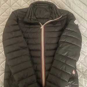 Moncler Daniel jacka - Säljer en stilren svart dunjacka från Moncler. Jackan har en dragkedja framtill Perfekt för kyligare dagar med sin lätta och bekväma design. Klassisk Moncler-logga på ärmen. Perfekt skick. Storlek 2