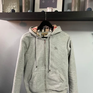 Grå hoodie från Burberry - Säljer en stilren grå hoodie från Burberry med dragkedja och broderad logga på bröstet. Insidan av huvan har det klassiska Burberry-mönstret. Säljer den i bra skick. Hör av dig vid intresse. 