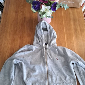 Grå hoodie från Ralph Lauren - Säljer en klassisk grå hoodie från Ralph Lauren med dragkedja och broderad logga på bröstet. Tröjan har en bekväm passform och är perfekt för en avslappnad stil. Den har en justerbar huva med snören och praktiska fickor framtill.