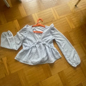 Ljusblå blus med prickar - Charmig ljusblå blus med små prickar och volangdetaljer. Blusen har långa ärmar och en v-ringad design. Perfekt för en söt och stilren look.