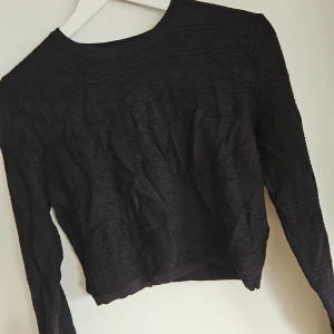 Svart långärmad tröja - Snygg svart tröja med långa ärmar och kort modell och ett subtilt mönster. Perfekt för en stilren look. Tröjan har en rund hals och är tillverkad i ett bekvämt material.