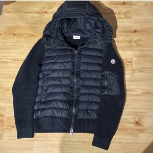 Svart pufferjacka från Moncler - Snygg svart pufferjacka från Moncler med huva och dragkedja. Jackan har en quiltad design på framsidan och ribbade ärmar. Perfekt för kyligare dagar. Köpt för 8000