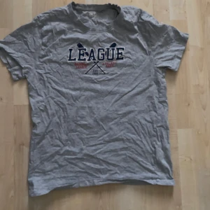 Grå t-shirt med tryck - Säljer en grå t-shirt med tryck av texten 'LEAGUE' . Perfekt för en avslappnad stil.