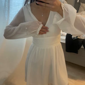 Vit klänning med knytband - Säljer en elegant vit klänning med v-ringning och långa ärmar. Klänningen har ett knytband i midjan och är perfekt för sommarens festligheter. Den har en luftig och lätt design som ger en romantisk känsla.