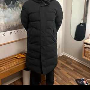 Svart lång dunjacka från elvine  - Säljer en svart lång dunjacka med huva. Jackan har en stilren design med dragkedja framtill och är perfekt för kalla dagar. Den är långärmad och har en pufferstil som ger extra värme och komfort. Nypris 4000 har haft den sen förra mars knappt använd.