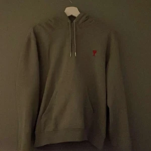 Ami Paris hoodie - Säljer en stilren grå hoodie med en liten röd detalj på bröstet. Tröjan har en klassisk design med huva och dragsko samt en stor ficka framtill. Perfekt för en avslappnad look.