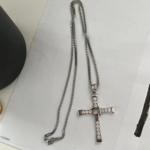 Silverfärgat halsband med korshänge - Elegant silverfärgat halsband med ett korshänge dekorerat med glittrande stenar. Perfekt för att ge en stilren touch till din outfit. Kedjan är av medellängd och har en klassisk låsning.