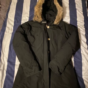 Svart parkajacka från Woolrich - Säljer en svart parkajacka från Woolrich med fuskpäls på huvan. Jackan har en quiltad insida och stängs med knappar. Perfekt för kalla dagar med sin varma och skyddande design. Pälsen går att ta av och sätta på. Pris går att diskutera! Hör av er vid frågor! 