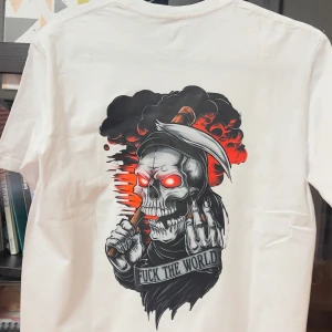 Vit t-shirt med dödskalletryck - Cool vit t-shirt med ett stort tryck av en dödskalle med glödande röda ögon och texten 'FUCK THE WORLD' på baksidan. Framsidan har texten 'THE REAPER' i gotisk stil. Perfekt för en edgy look.